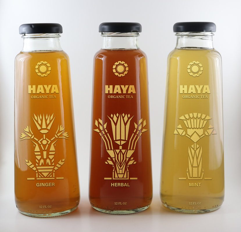 Three Haya Organic Tea bottles with gold Egyptian floral label designs in ginger, herbal, and mint flavors
